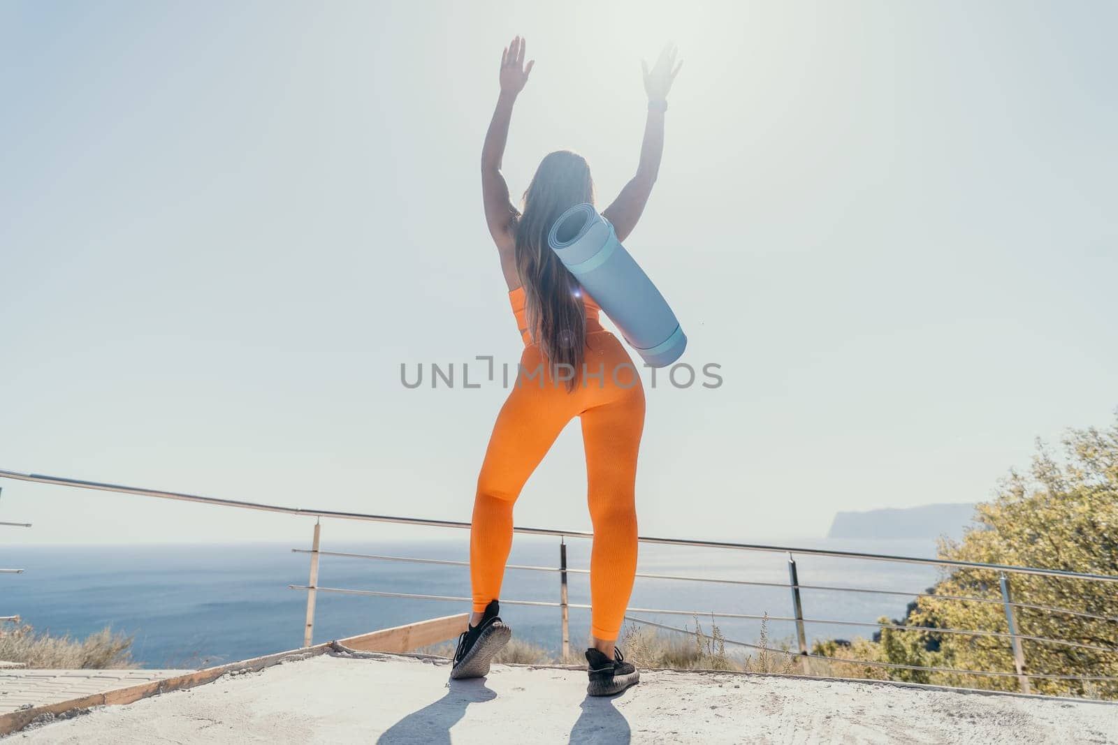 A young woman in orange yoga clothes with a blue mat slung over her shoulder raises her arms in triumph against a bright blue sky.