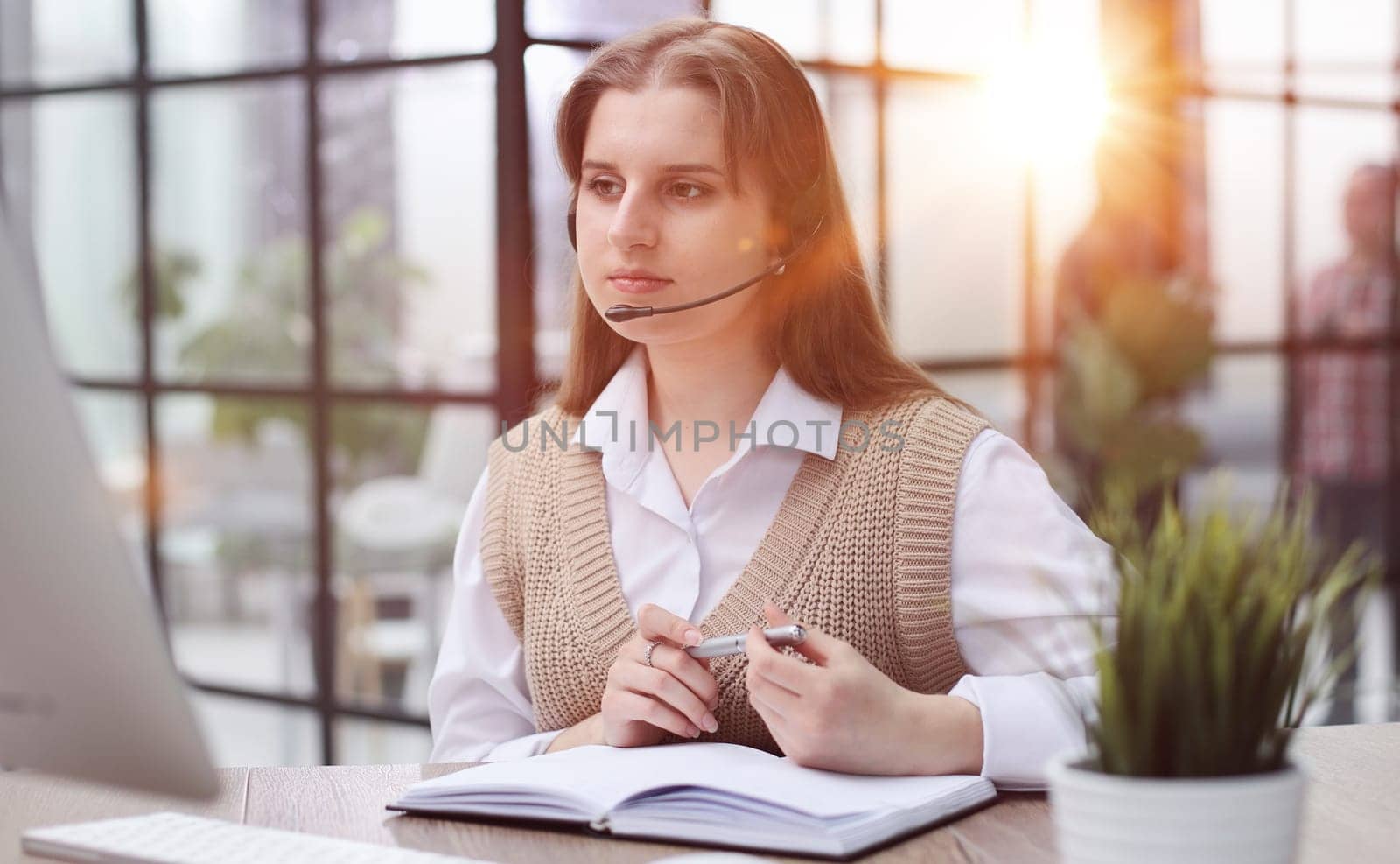 Attractive confident business woman, office manager, wearing headset using laptop while making, writing financial report, using pen, on paper working indoors.