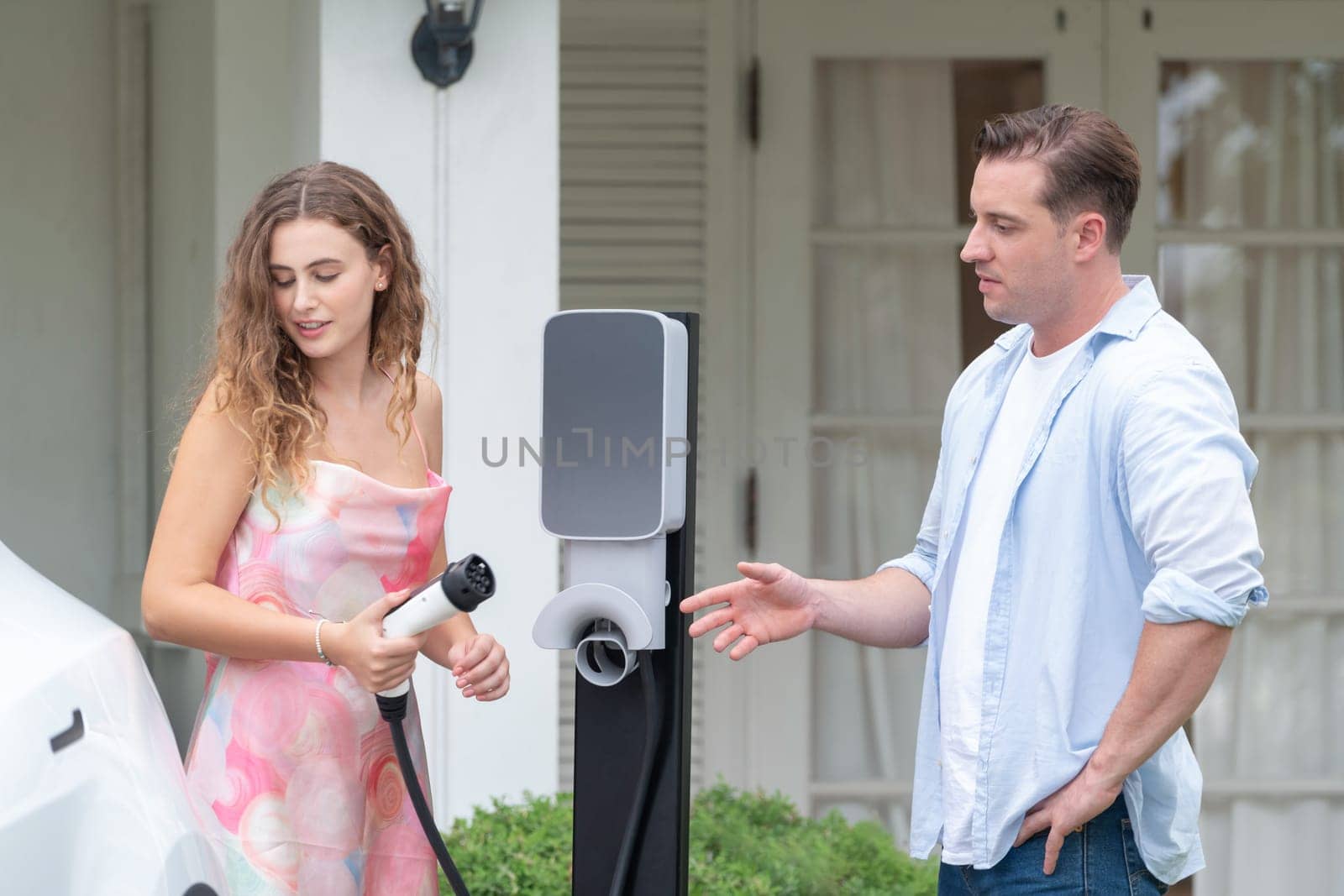 Happy and lovely couple with eco-friendly conscious recharging electric vehicle from EV home charging station. EV car technology utilized for residential home to future sustainability. Synchronos