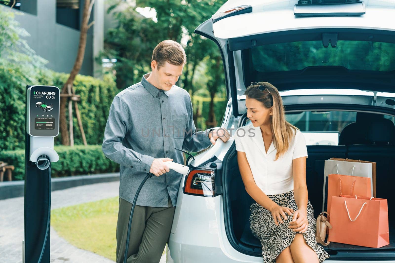 Young couple travel with EV electric car charging in green sustainable city outdoor garden in summer shows urban sustainability lifestyle by green clean rechargeable energy of electric vehicle innards