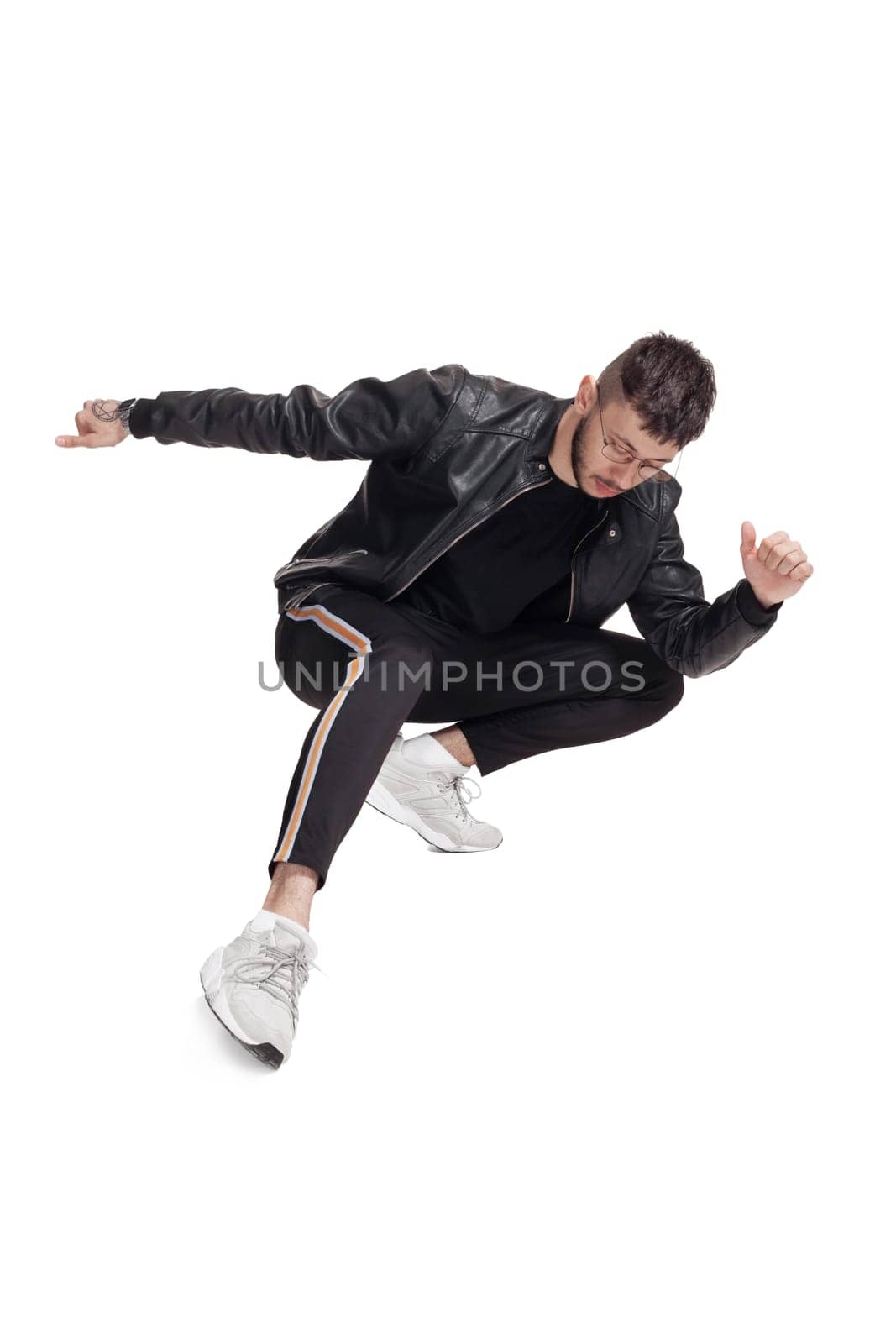 Full-length photo of a modern performer in glasses, black leather jacket, t-shirt, sports pants and light sneakers fooling around in studio. Indoor photo of a good-looking fellow dancing isolated on white background. Music and imagination.
