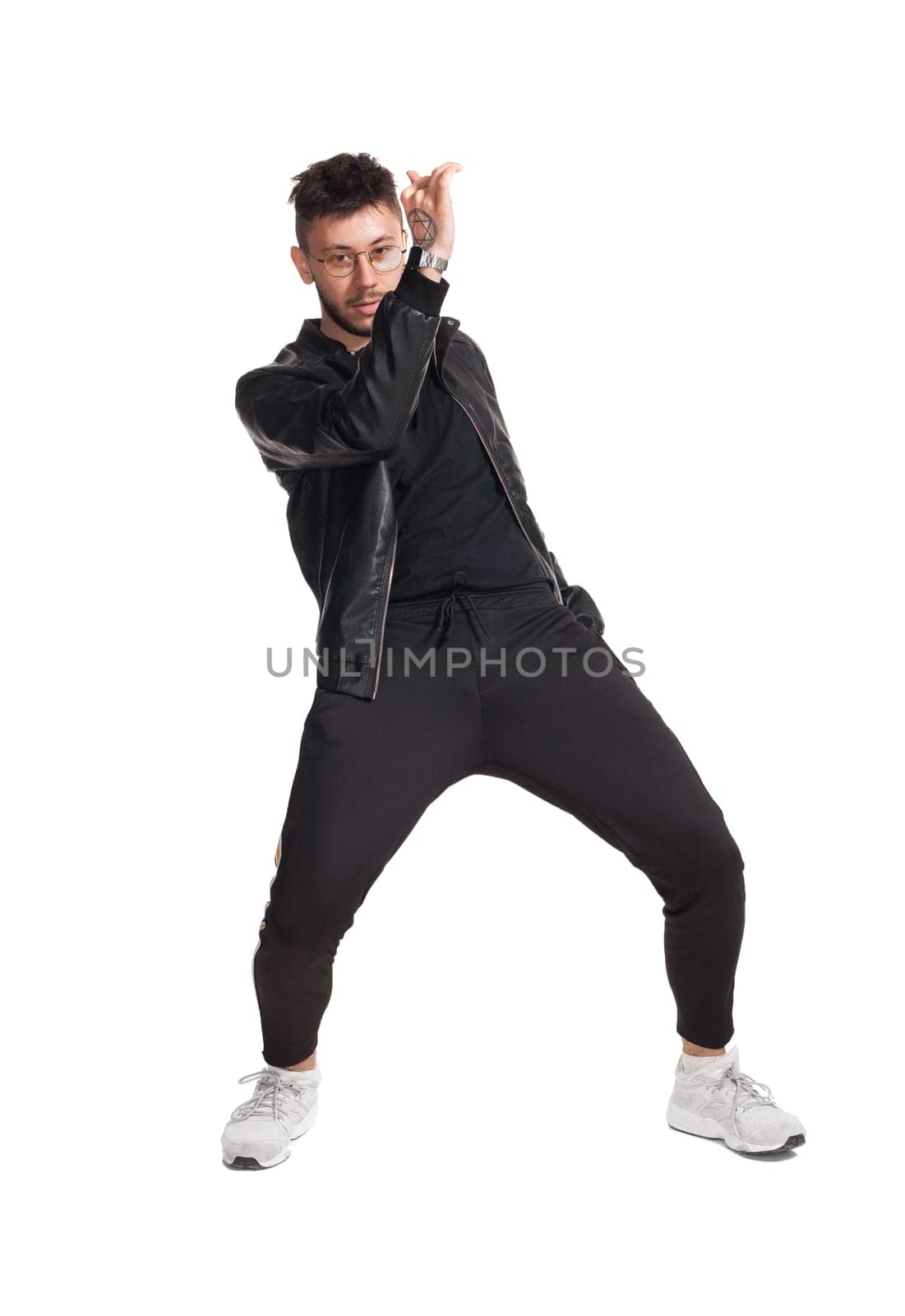 Full-length photo of a modern performer in glasses, black leather jacket, t-shirt, sports pants and light sneakers fooling around in studio. Indoor photo of a good-looking man dancing and looking at the camera isolated on white background. Music and imagination.