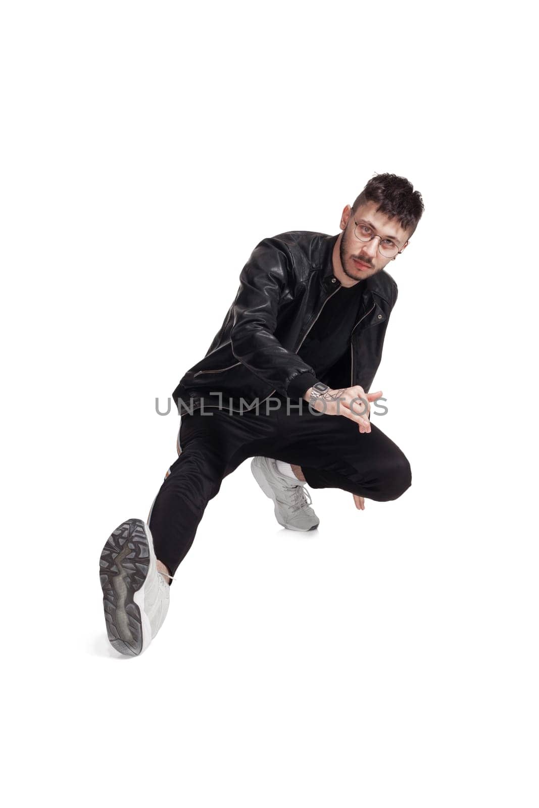 Full-length photo of a modern performer in glasses, black leather jacket, t-shirt, sports pants and light sneakers fooling around in studio. Indoor photo of a good-looking male dancing sitting isolated on white background. Music and imagination.