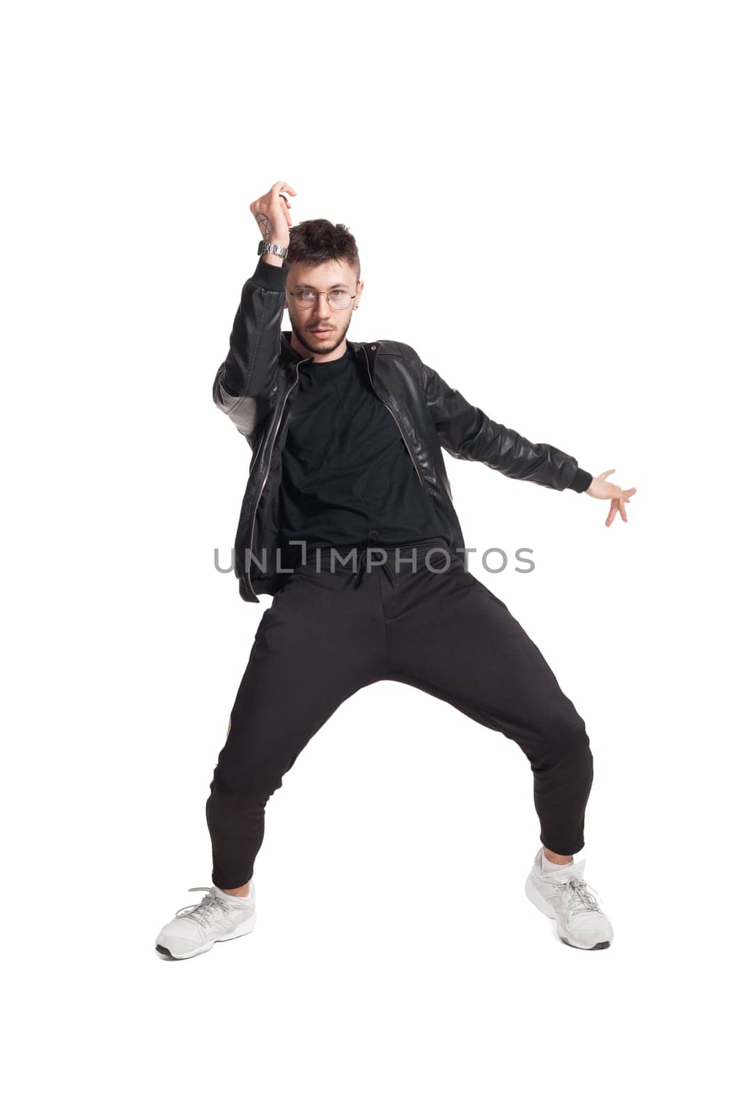 Full-length photo of a modern performer in glasses, black leather jacket, t-shirt, sports pants and light sneakers fooling around in studio. Indoor photo of an attractive fellow dancing and looking at the camera isolated on white background. Music and imagination.