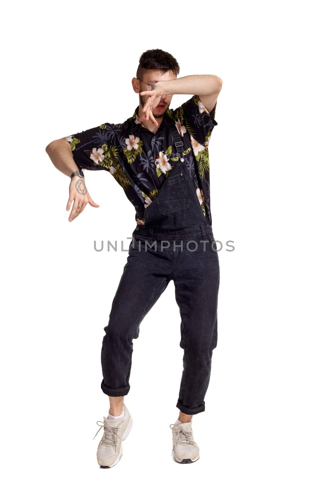 Full-length portrait of a cool man in glasses, black jumpsuit, colorful t-shirt and gray sneakers fooling around in studio. Indoor photo of a man dancing isolated on white background. Music and imagination. Copy space.