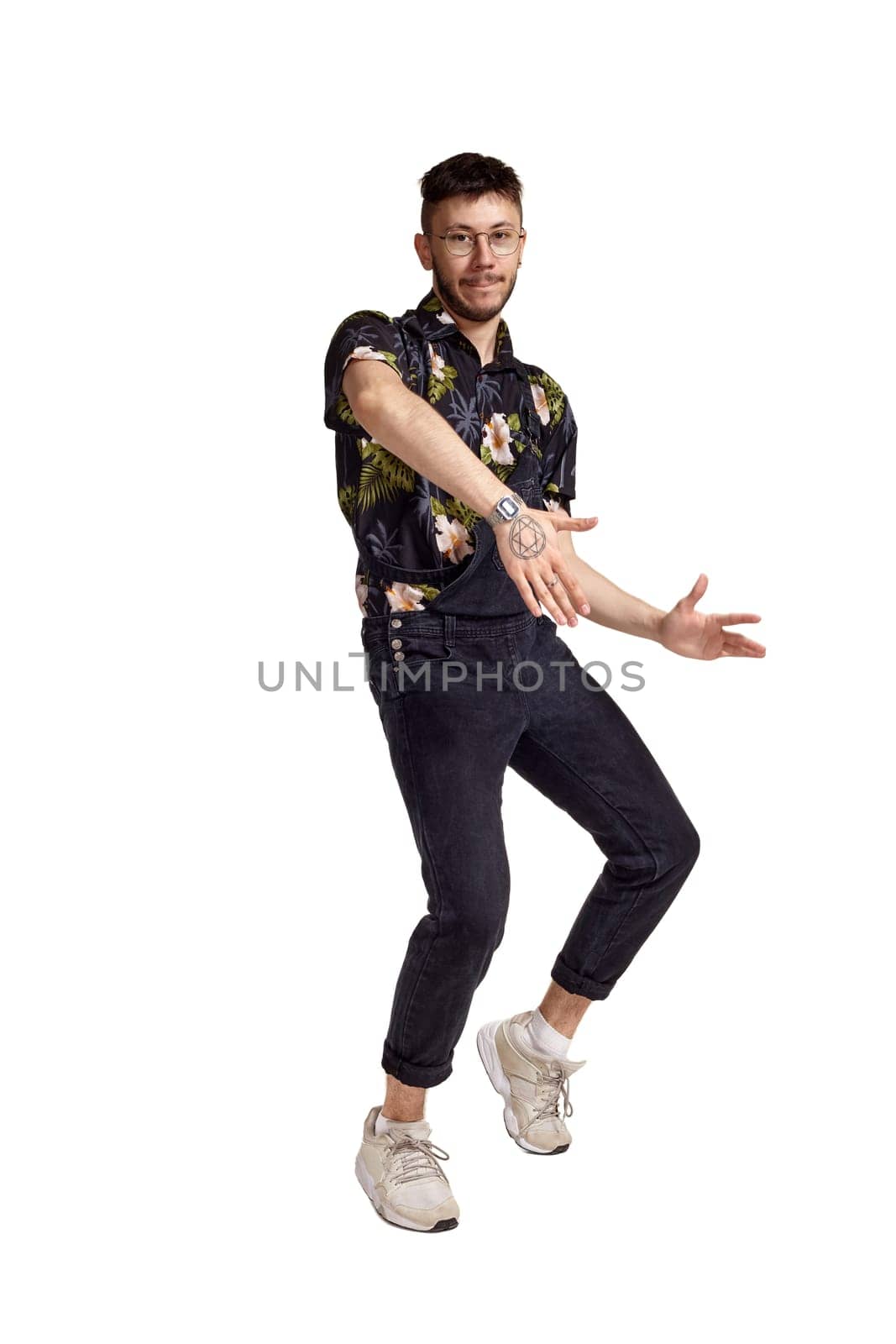 Full-length portrait of a cool guy in glasses, black jumpsuit, colorful t-shirt and gray sneakers fooling around in studio. Indoor photo of a man dancing isolated on white background. Music and imagination.