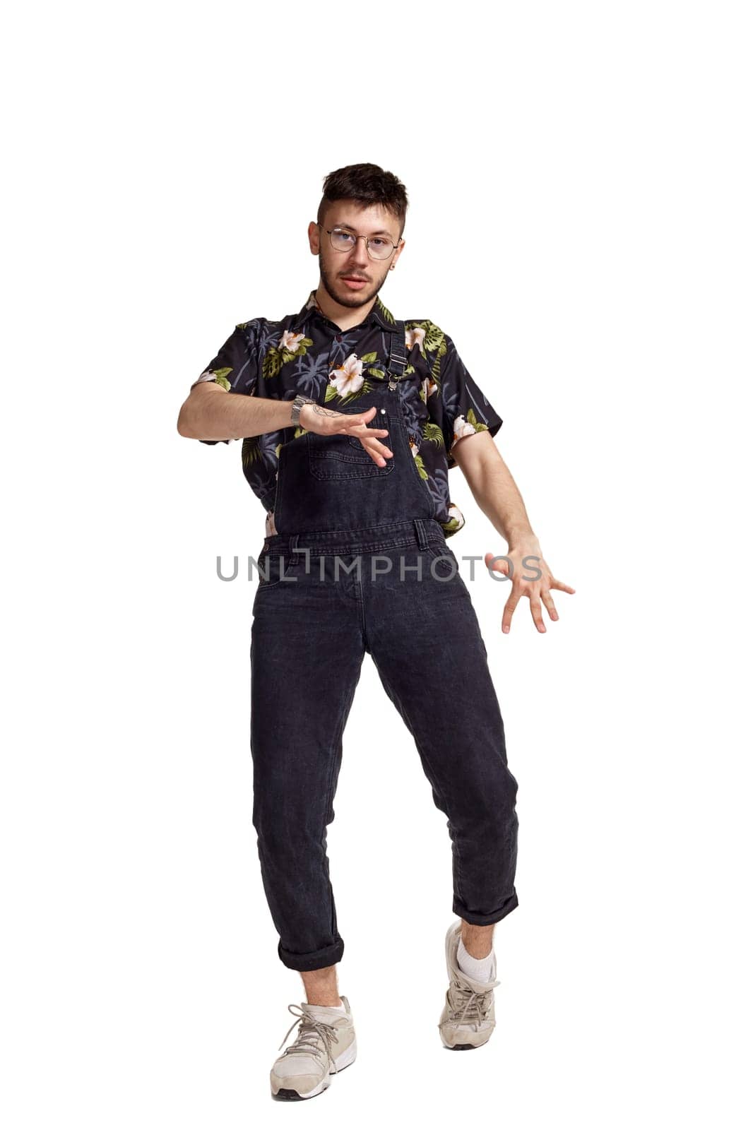 Full-length portrait of a fashionable dancer in glasses, black jumpsuit, colorful t-shirt and gray sneakers fooling around in studio. Indoor photo of a man dancing isolated on white background. Music and imagination. Copy space.