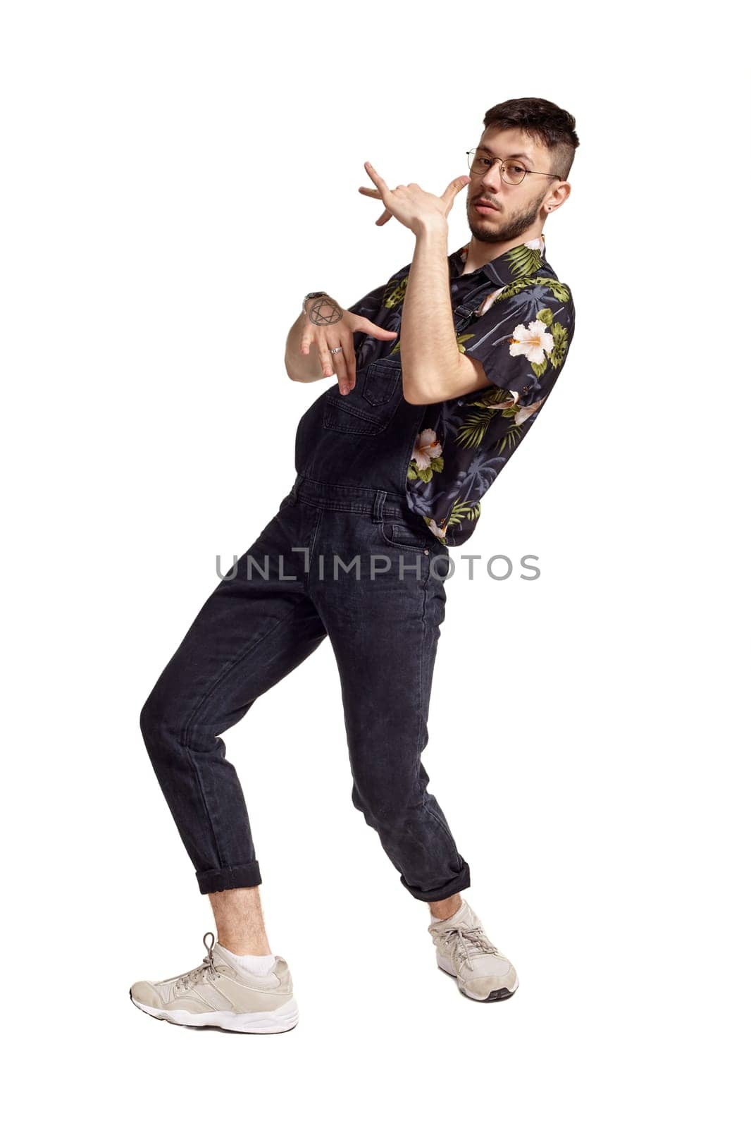 Full-length portrait of a graceful dancer in glasses, black jumpsuit, colorful t-shirt and gray sneakers fooling around in studio. Indoor photo of a man dancing sideways isolated on white background. Music and imagination.