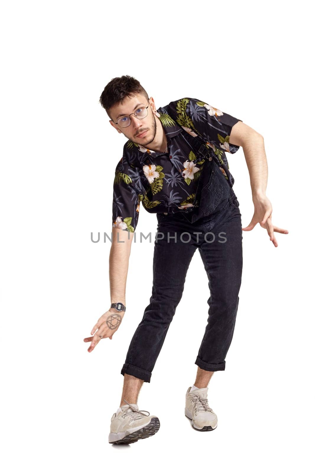 Full-length portrait of a graceful person in glasses, black jumpsuit, colorful t-shirt and gray sneakers fooling around in studio. Indoor photo of a man dancing isolated on white background. Music and imagination. Copy space.