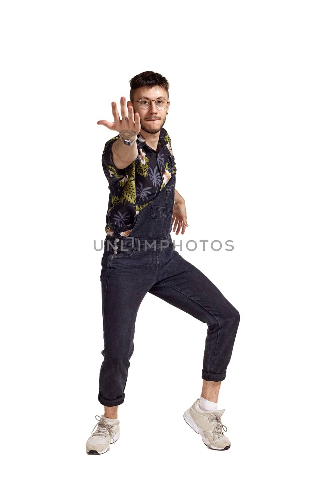 Full-length portrait of a graceful man in glasses, black jumpsuit, colorful t-shirt and gray sneakers fooling around in studio. Indoor photo of a man dancing isolated on white background. Music and imagination concept.