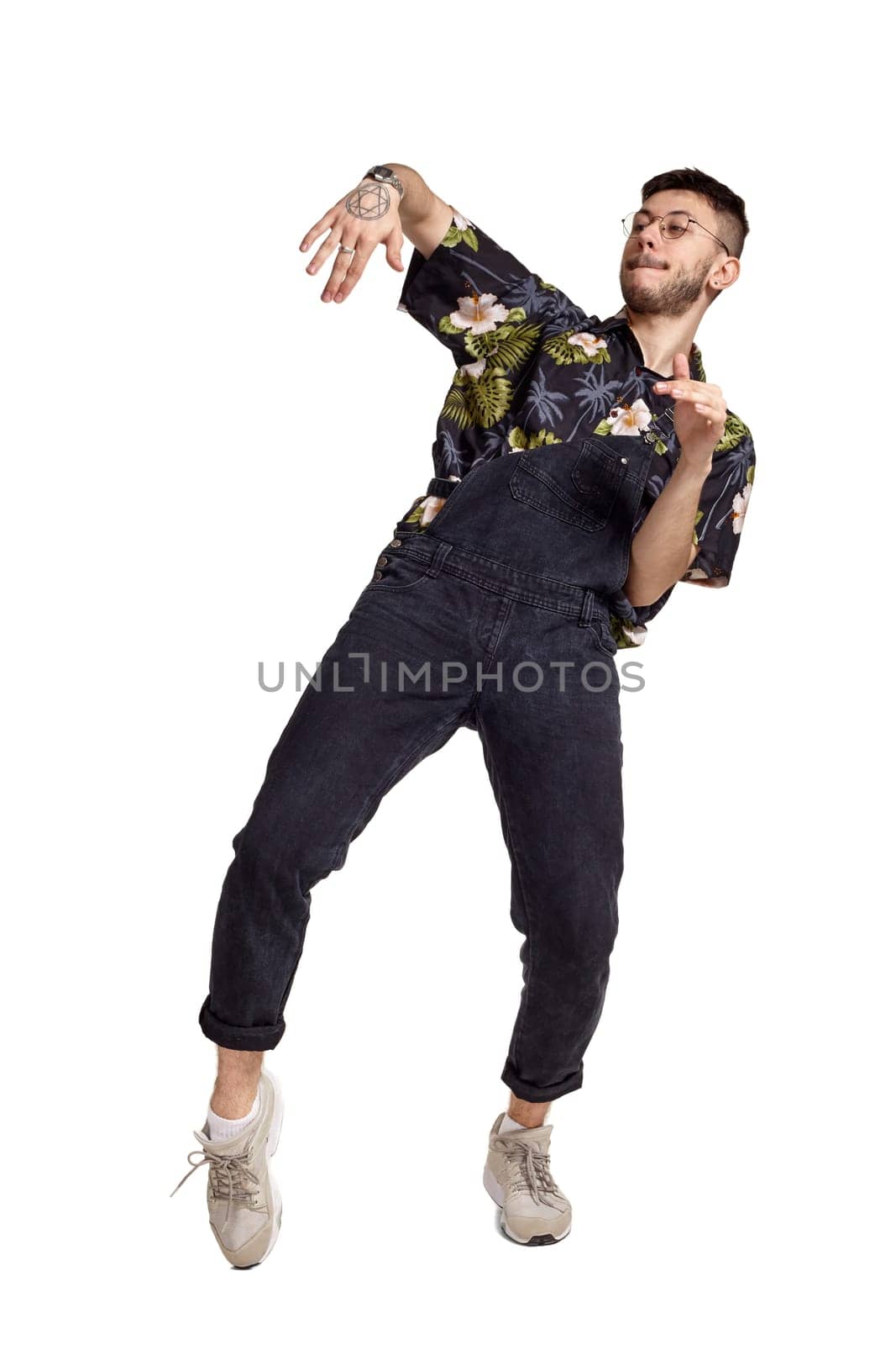 Full-length portrait of an athletic person in glasses, black jumpsuit, colorful t-shirt and gray sneakers fooling around in studio. Indoor photo of a man dancing isolated on white background. Music and imagination concept.