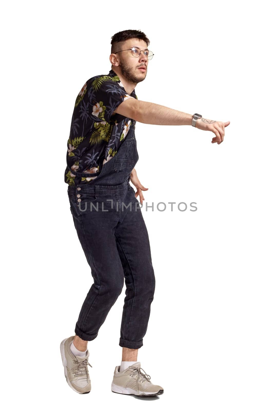 Full-length portrait of an athletic male in glasses, black jumpsuit, colorful t-shirt and gray sneakers fooling around in studio. Indoor photo of a man dancing isolated on white background. Music and imagination. Copy space.