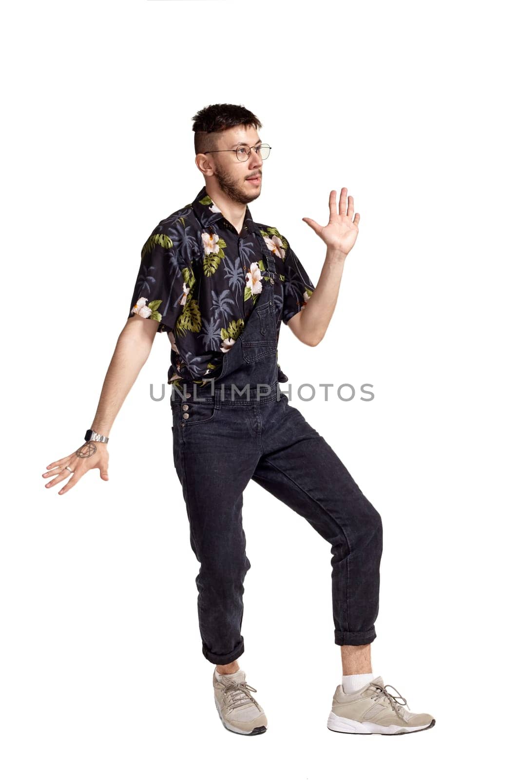 Full-length portrait of an athletic man in glasses, black jumpsuit, colorful t-shirt and gray sneakers fooling around in studio. Indoor photo of a man dancing sideways isolated on white background. Music and imagination.