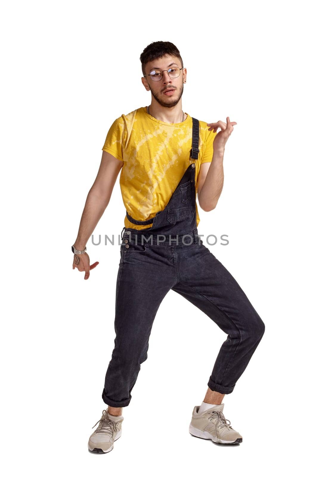 Full-length portrait of a fasionable man in glasses, black jumpsuit, yellow t-shirt and gray sneakers fooling around in studio. Indoor photo of a man making dancing elements isolated on white background. Music and imagination. Copy space.