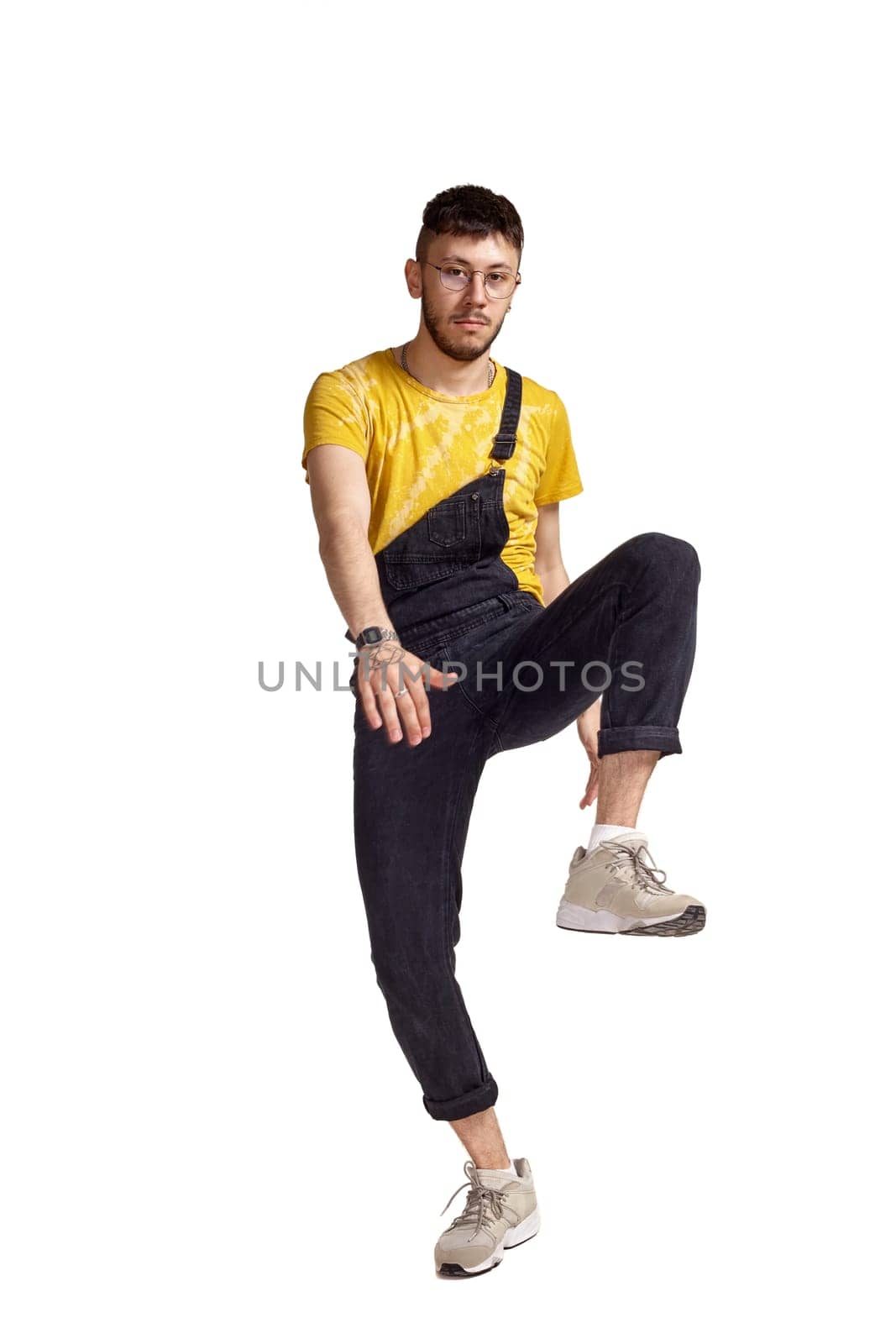 Full-length portrait of a stylish dancer in glasses, black jumpsuit, yellow t-shirt and gray sneakers fooling around in studio. Indoor photo of a man making dancing elements raising his leg isolated on white background. Music and imagination.
