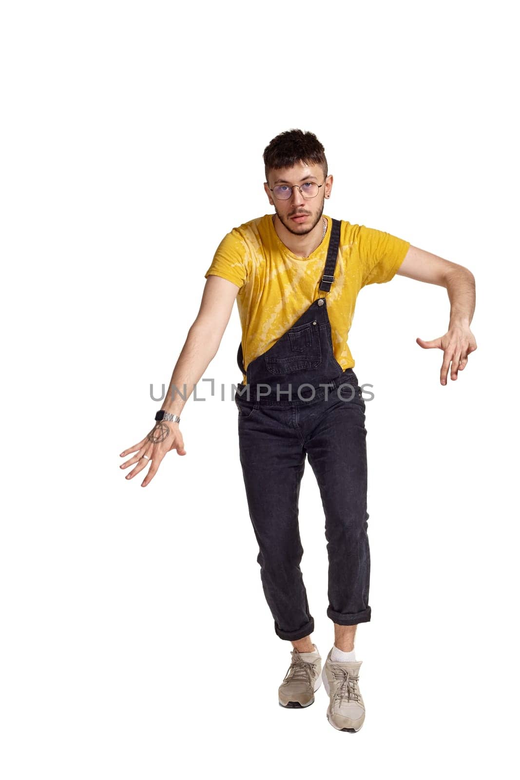 Full-length portrait of a stylish male in glasses, black jumpsuit, yellow t-shirt and gray sneakers fooling around in studio. Indoor photo of a man making dancing elements isolated on white background. Music and imagination concept.