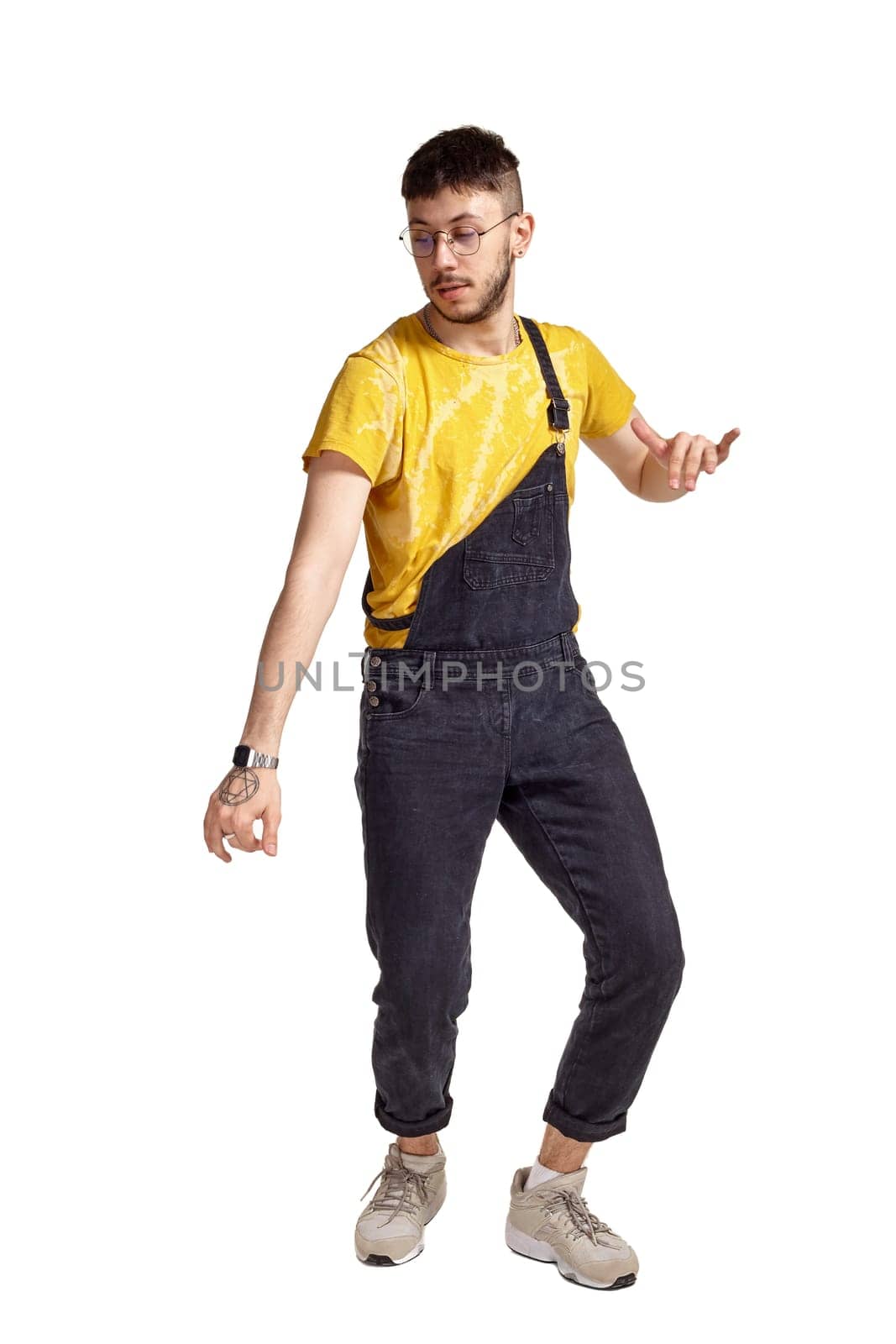Full-length portrait of a stylish guy in glasses, black jumpsuit, yellow t-shirt and gray sneakers fooling around in studio. Indoor photo of a man making dancing elements isolated on white background and looking down. Music and imagination.