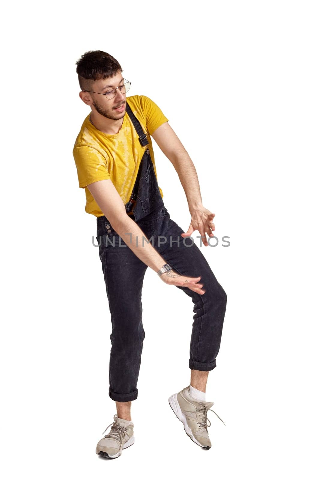 Full-length portrait of a sporty person in glasses, black jumpsuit, yellow t-shirt and gray sneakers fooling around in studio. Indoor photo of a man making dancing elements isolated on white background. Music and imagination.