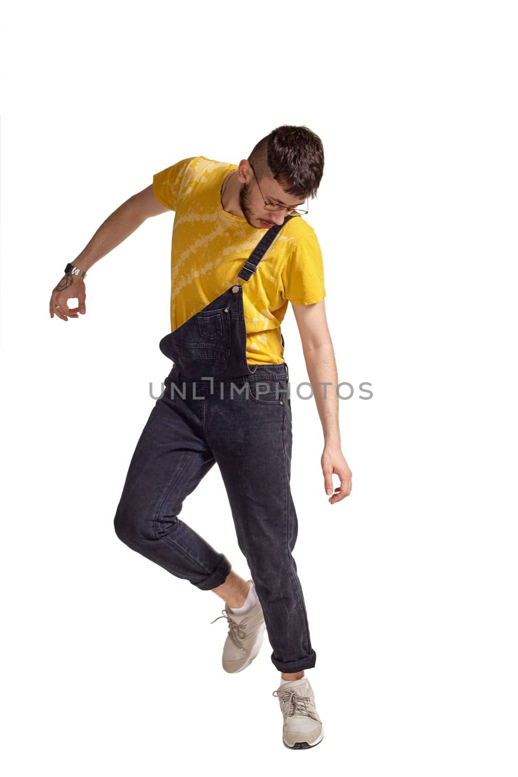Full-length portrait of an athletic male in glasses, black jumpsuit, yellow t-shirt and gray sneakers fooling around in studio. Indoor photo of a man making dancing elements isolated on white background and looking down. Music and imagination.