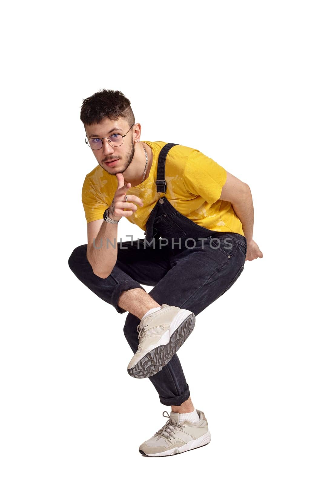 Full-length portrait of a handsome dancer in glasses, black jumpsuit, yellow t-shirt and gray sneakers fooling around in studio. Indoor photo of a man making dancing elements isolated on white background and looking at the camera. Music and imagination.