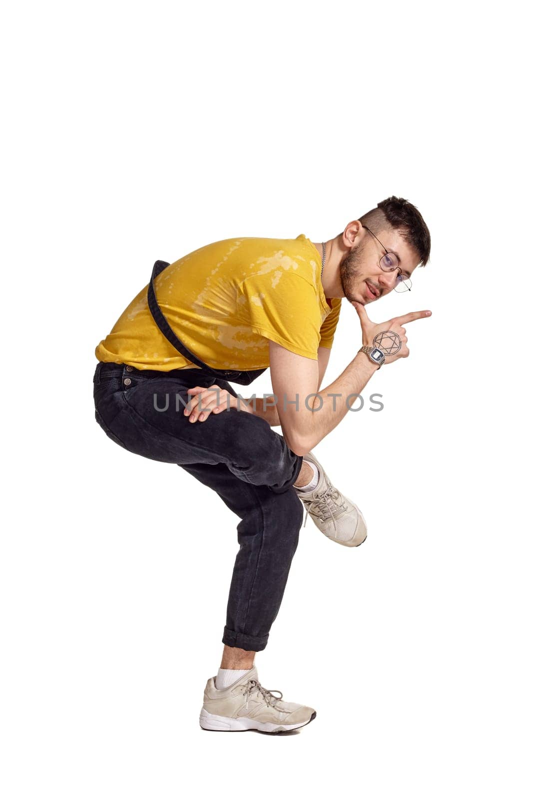 Full-length portrait of a handsome male in glasses, black jumpsuit, yellow t-shirt and gray sneakers fooling around in studio. Indoor photo of a man making dancing elements isolated on white background and looking at the camera. Music and imagination.