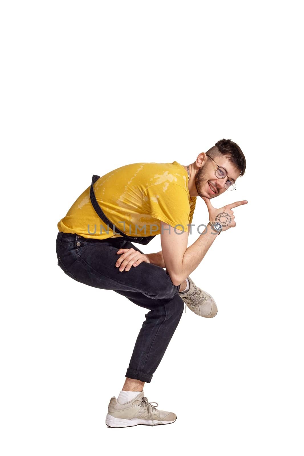Full-length portrait of a handsome guy in glasses, black jumpsuit, yellow t-shirt and gray sneakers fooling around in studio. Indoor photo of a man making dancing elements sitting isolated on white background. Music and imagination.