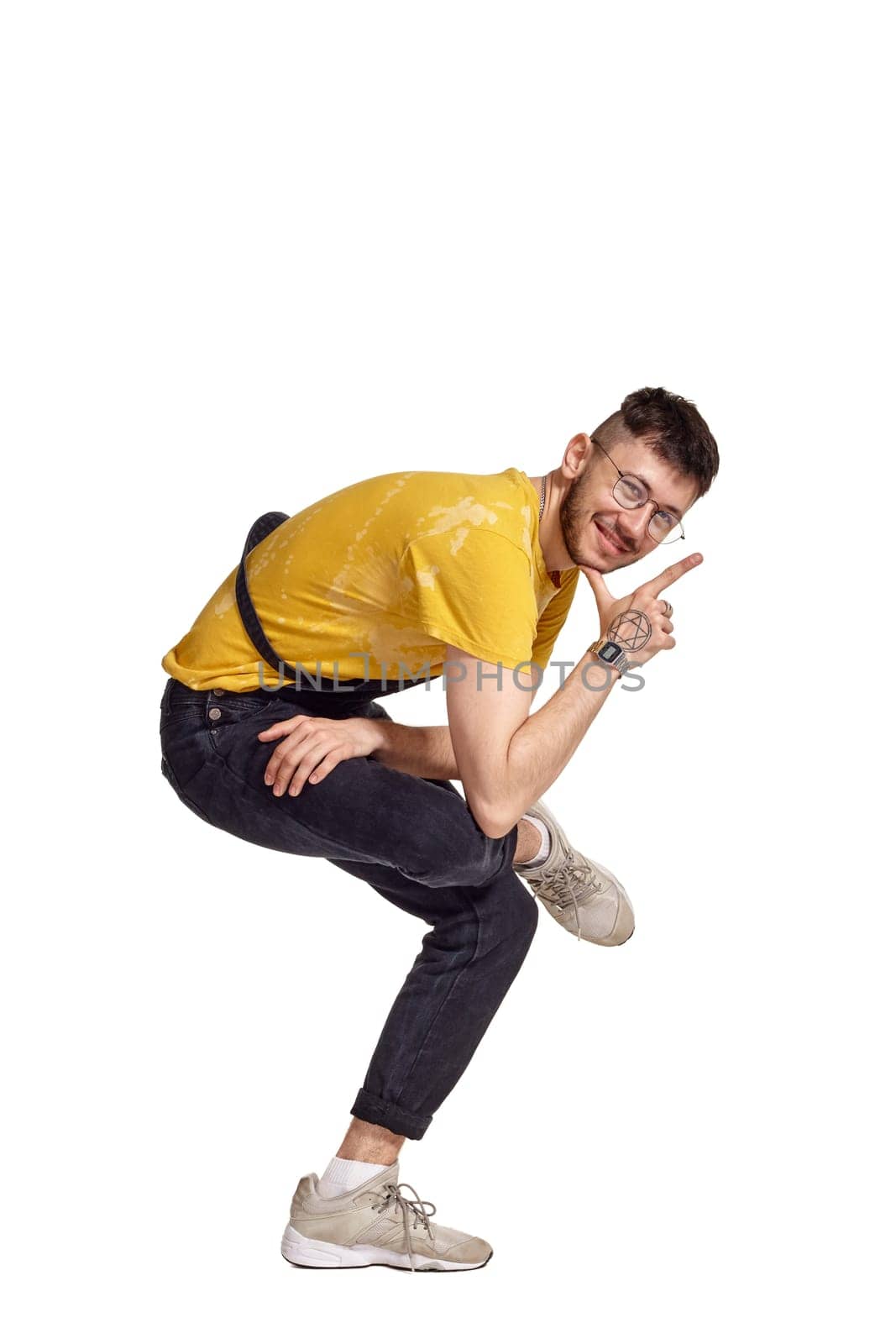 Full-length portrait of a funny dancer in glasses, black jumpsuit, yellow t-shirt and gray sneakers fooling around in studio. Indoor photo of a man making dancing elements isolated on white background. Music and imagination.