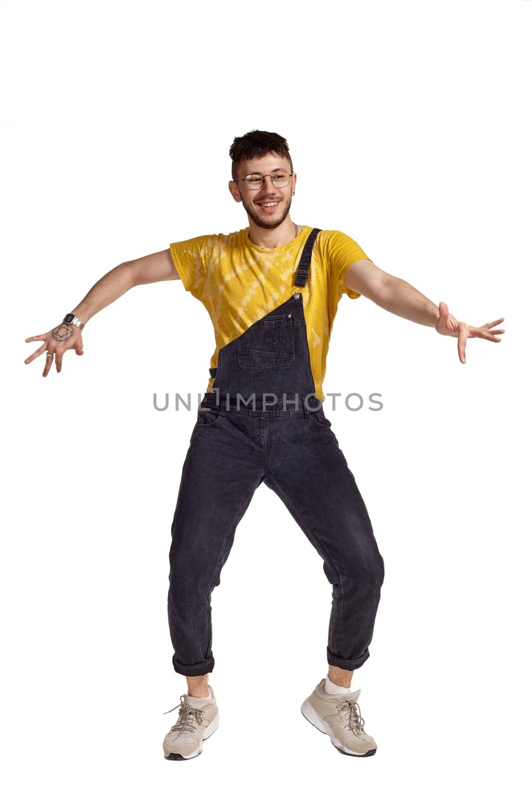 Full-length portrait of a funny man in glasses, black jumpsuit, yellow t-shirt and gray sneakers fooling around in studio. Indoor photo of a man making dancing elements isolated on white background. Music and imagination.