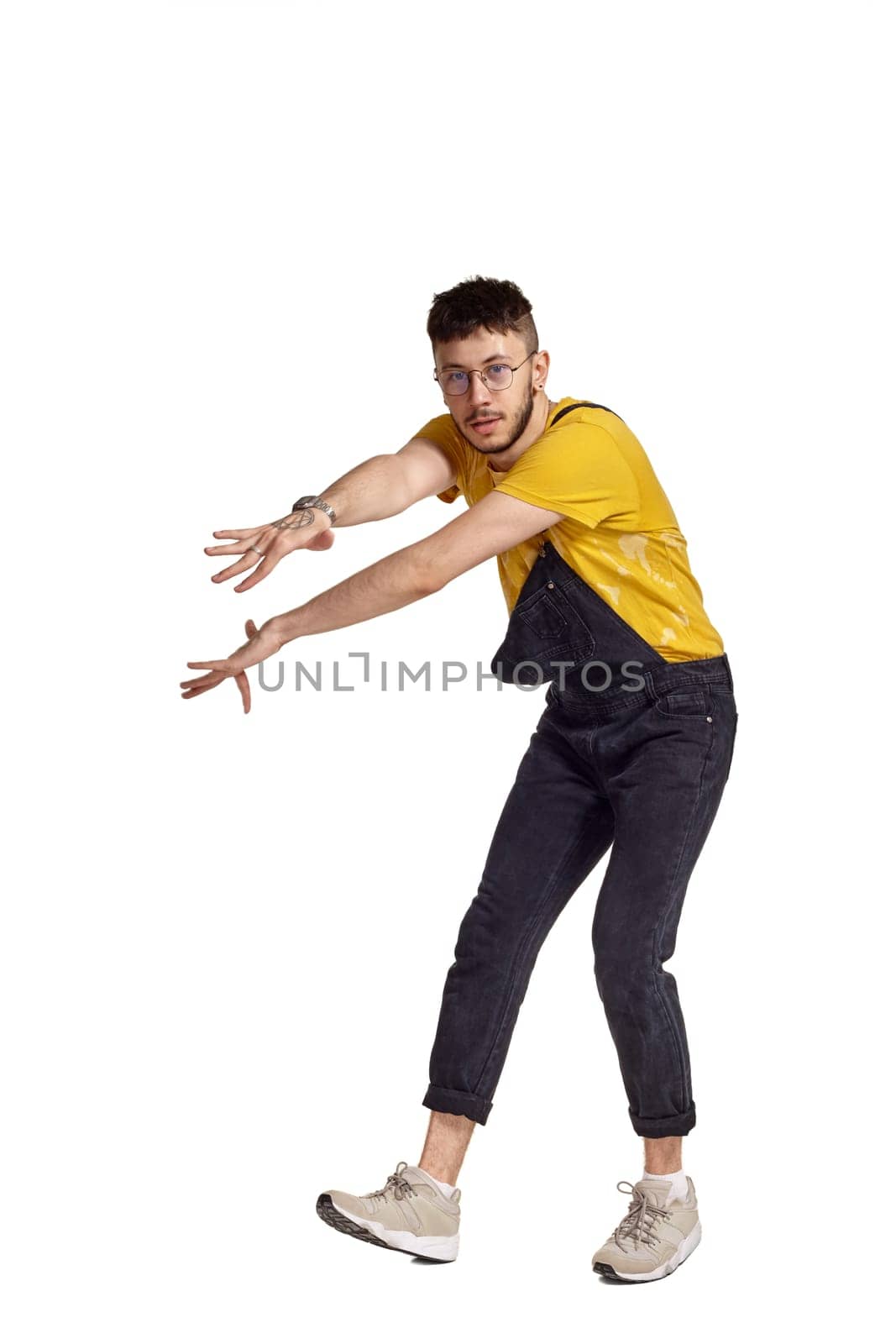 Full-length portrait of a graceful guy in glasses, black jumpsuit, yellow t-shirt and gray sneakers fooling around in studio. Indoor photo of a man making dancing elements isolated on white background. Music and imagination.