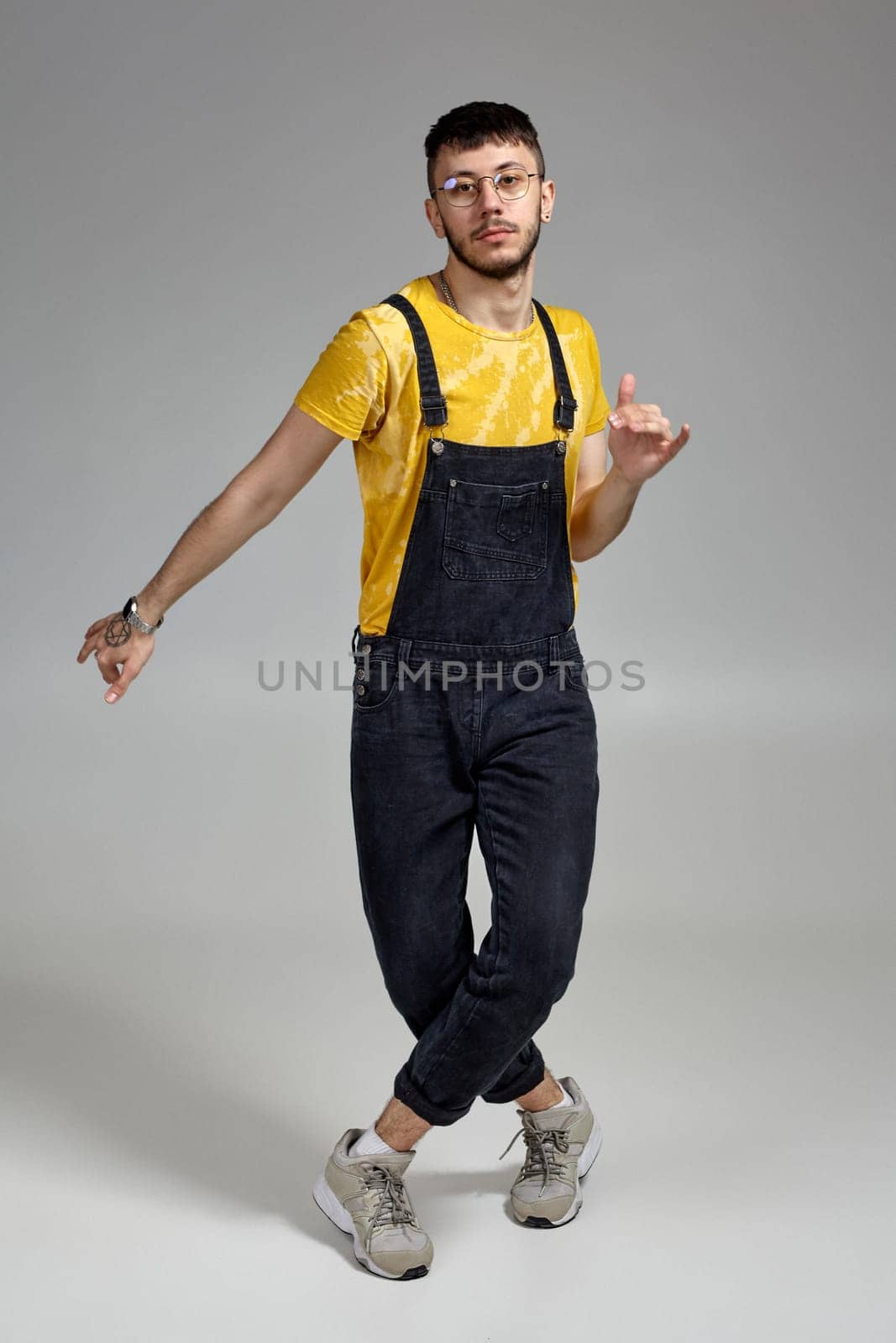 Full-length portrait of a stylish man in glasses, black jumpsuit, yellow t-shirt and gray sneakers fooling around in studio. Indoor photo of a man dancing on a gray background. Music and imagination concept.