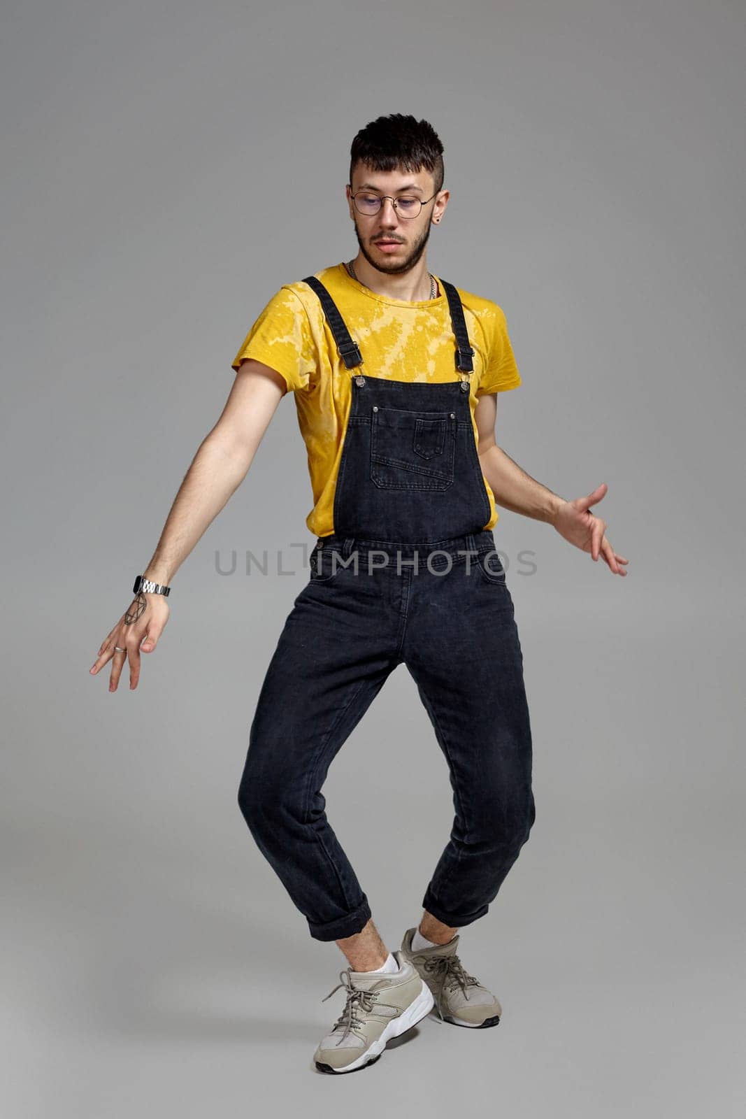 Full-length portrait of a stylish male in glasses, black jumpsuit, yellow t-shirt and gray sneakers fooling around in studio. Indoor photo of a man dancing on a gray background. Music and imagination.