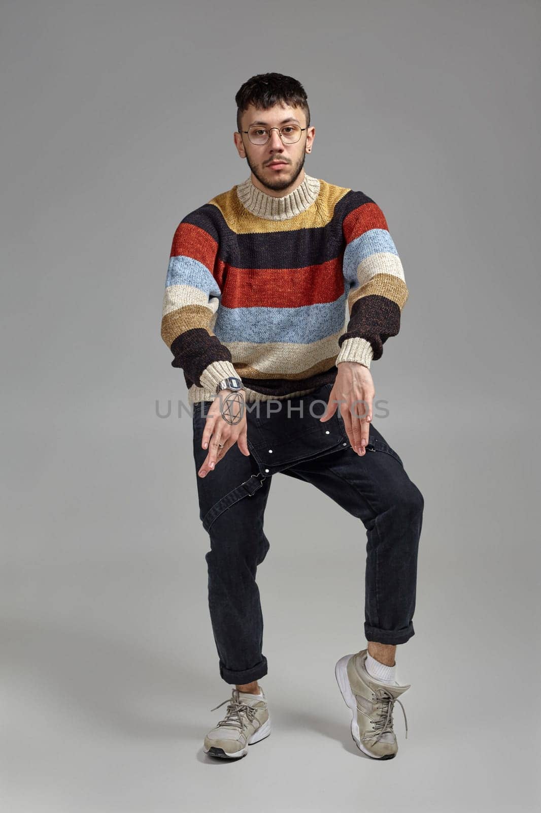 Full-length portrait of an athletic male in glasses, black jumpsuit, multi-colored sweater and gray sneakers fooling around in studio. Indoor photo of a man dancing on a gray background. Music and imagination.