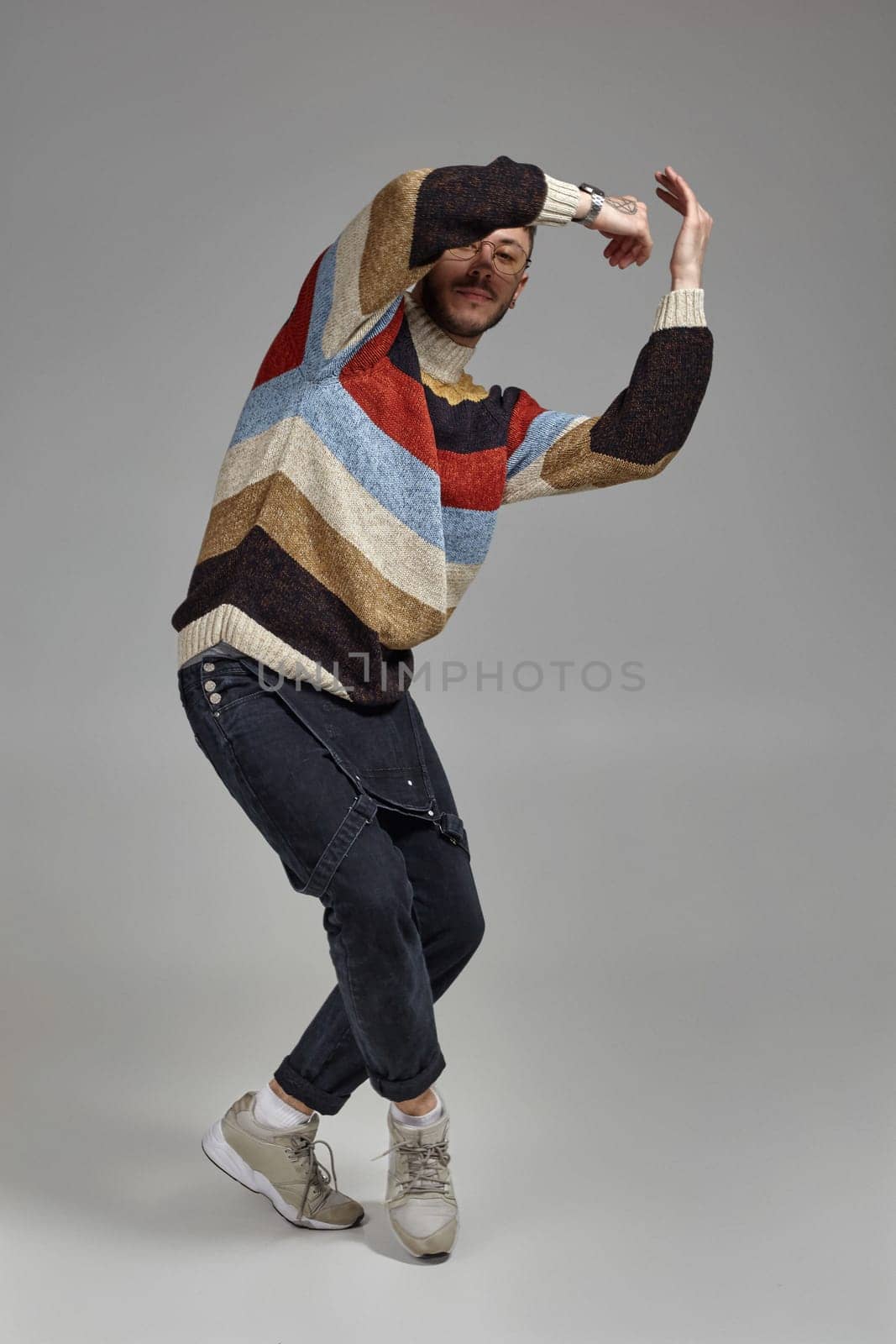Full-length portrait of a good-looking male in glasses, black jumpsuit, multi-colored sweater and gray sneakers fooling around in studio. Indoor photo of a man dancing raising his hands on a gray background. Music and imagination.