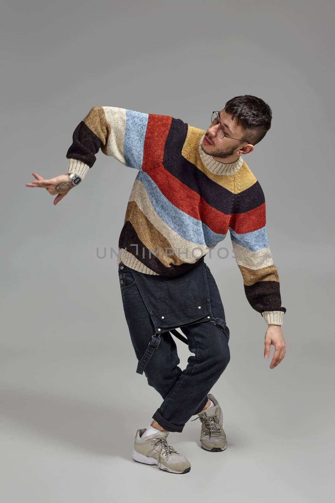 Full-length portrait of a funny dancer in glasses, black jumpsuit, multi-colored sweater and gray sneakers fooling around in studio. Indoor photo of a guy making dance elements on a gray background. Music and imagination.