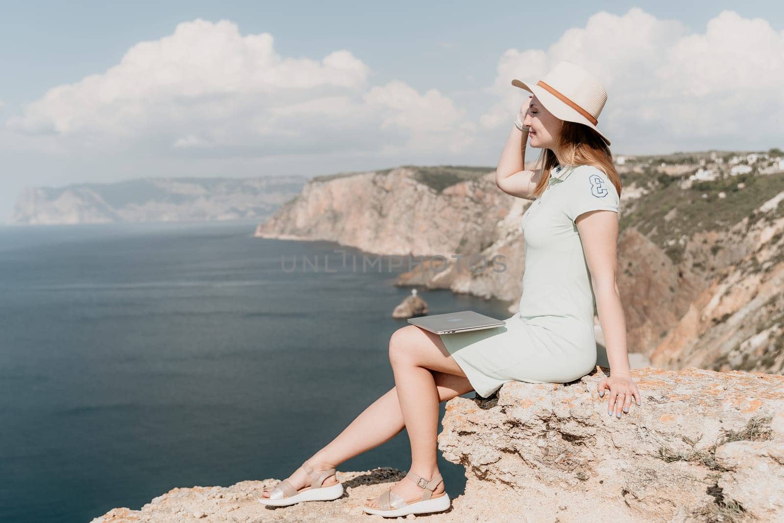 A woman in a straw hat sits on a cliff, holding a laptop, overlooking the vast blue ocean and rocky coastline.