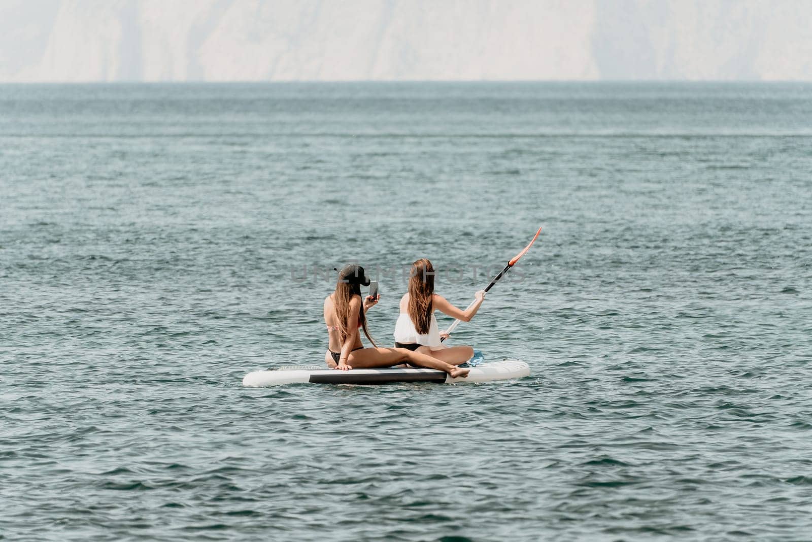 Two women are on a surfboard in the ocean. One of them is wearing a white shirt and the other is wearing a black bikini