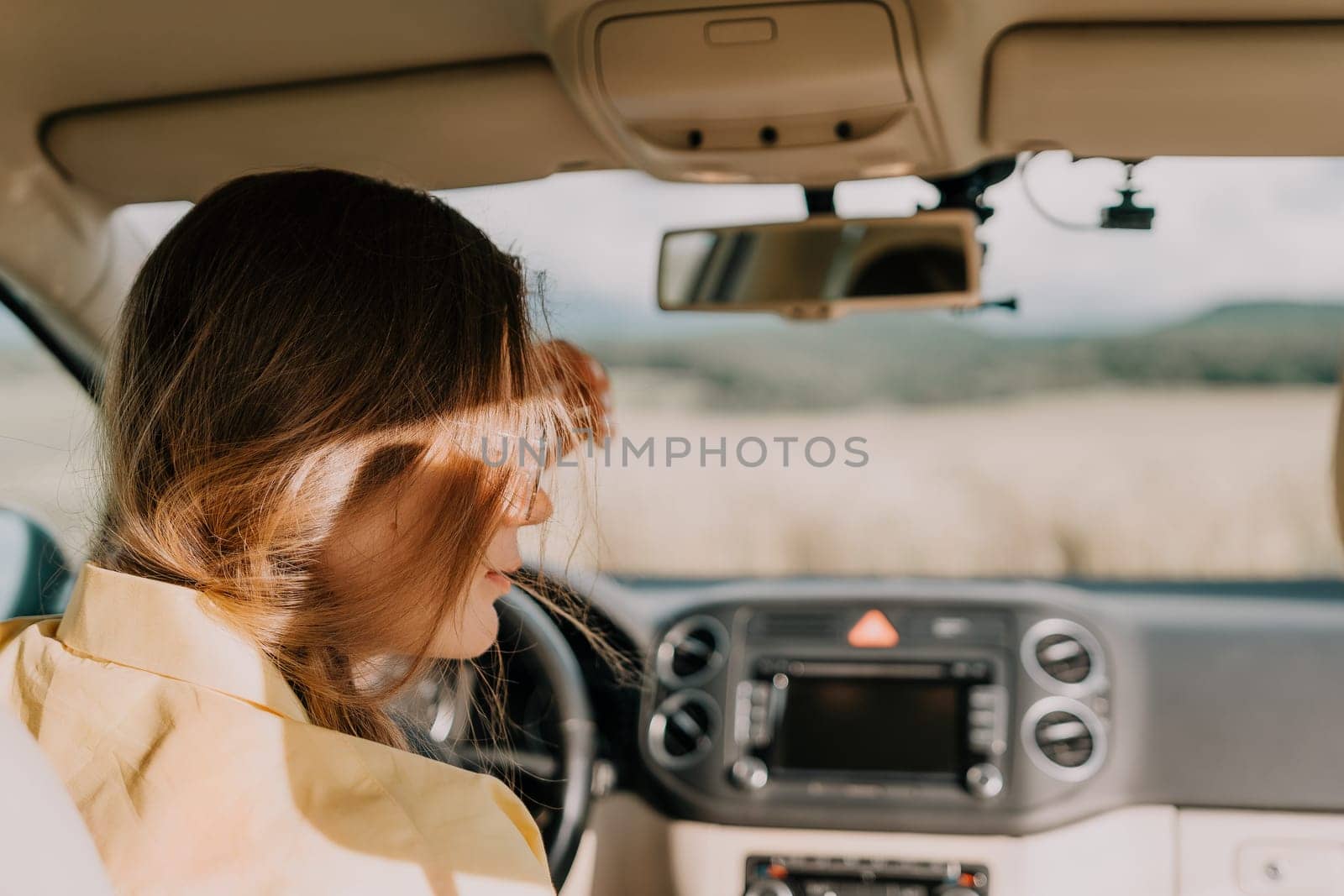 A young woman driving a car with her long brown hair blowing in the wind on a sunny day.