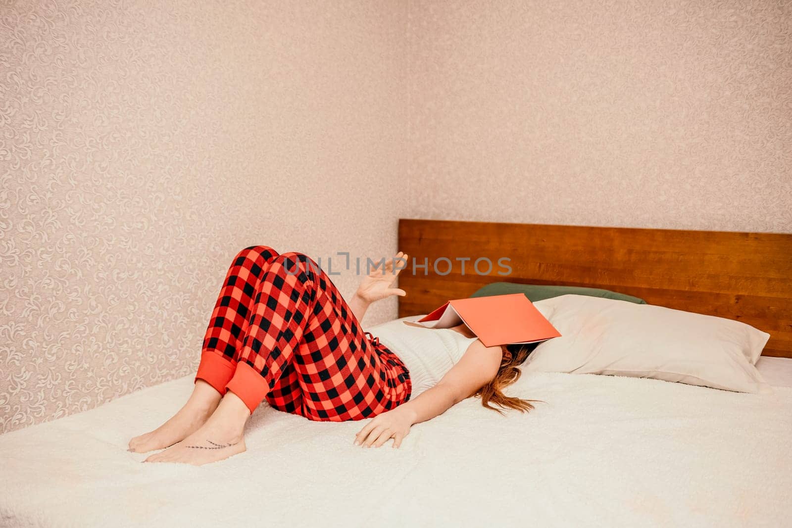 Young brunette woman works and studies from home on a laptop, while lying on her bed in red checkered pants. Balancing work and studies at home