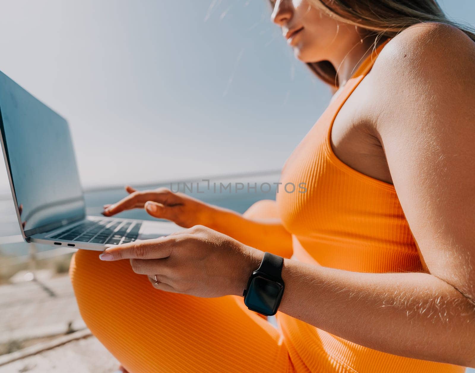 A woman wearing an orange outfit is sitting outdoors, using a laptop. She is likely working or browsing the internet.