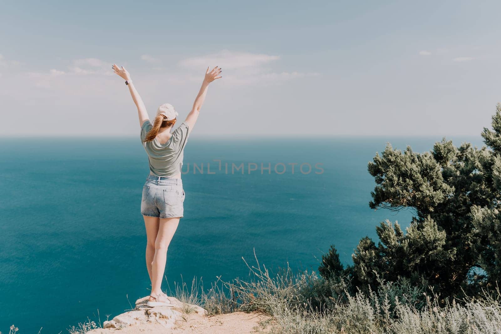 A woman with her arms raised in the air stands on a cliff looking out at the deep blue sea, capturing a sense of liberation and joy.