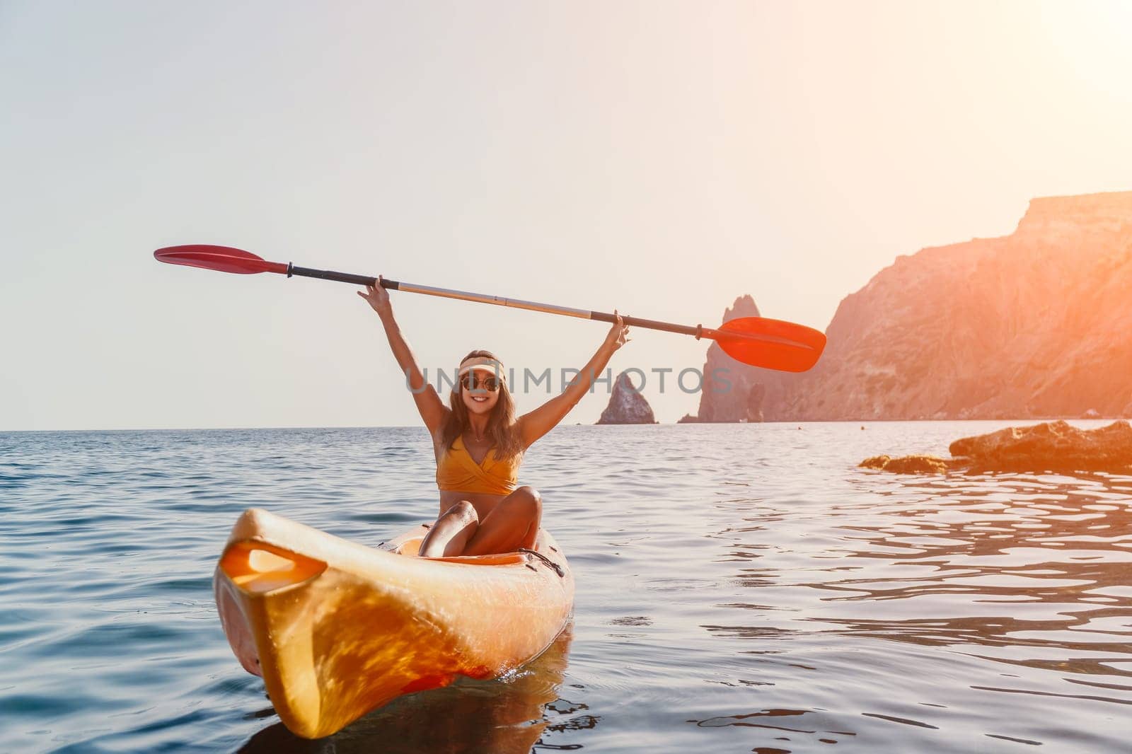 A woman in a bikini paddles a kayak with raised arms, enjoying a scenic ocean view with mountains in the distance.