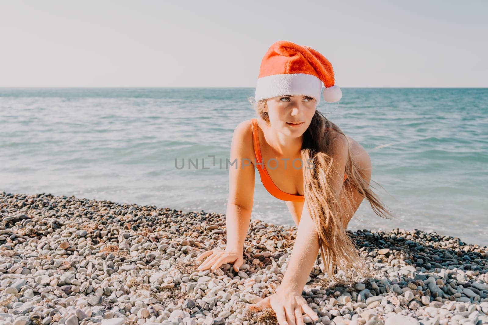 A woman wearing a red Santa hat and a bright orange bikini crawls along a pebble beach by the sea.