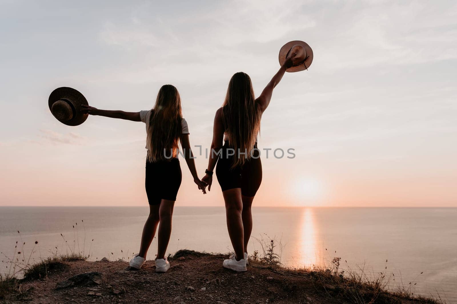 Two women in black shorts and white sneakers stand with their backs to the camera on a cliff, holding hands. They face the ocean at sunset and are silhouetted against the sky.