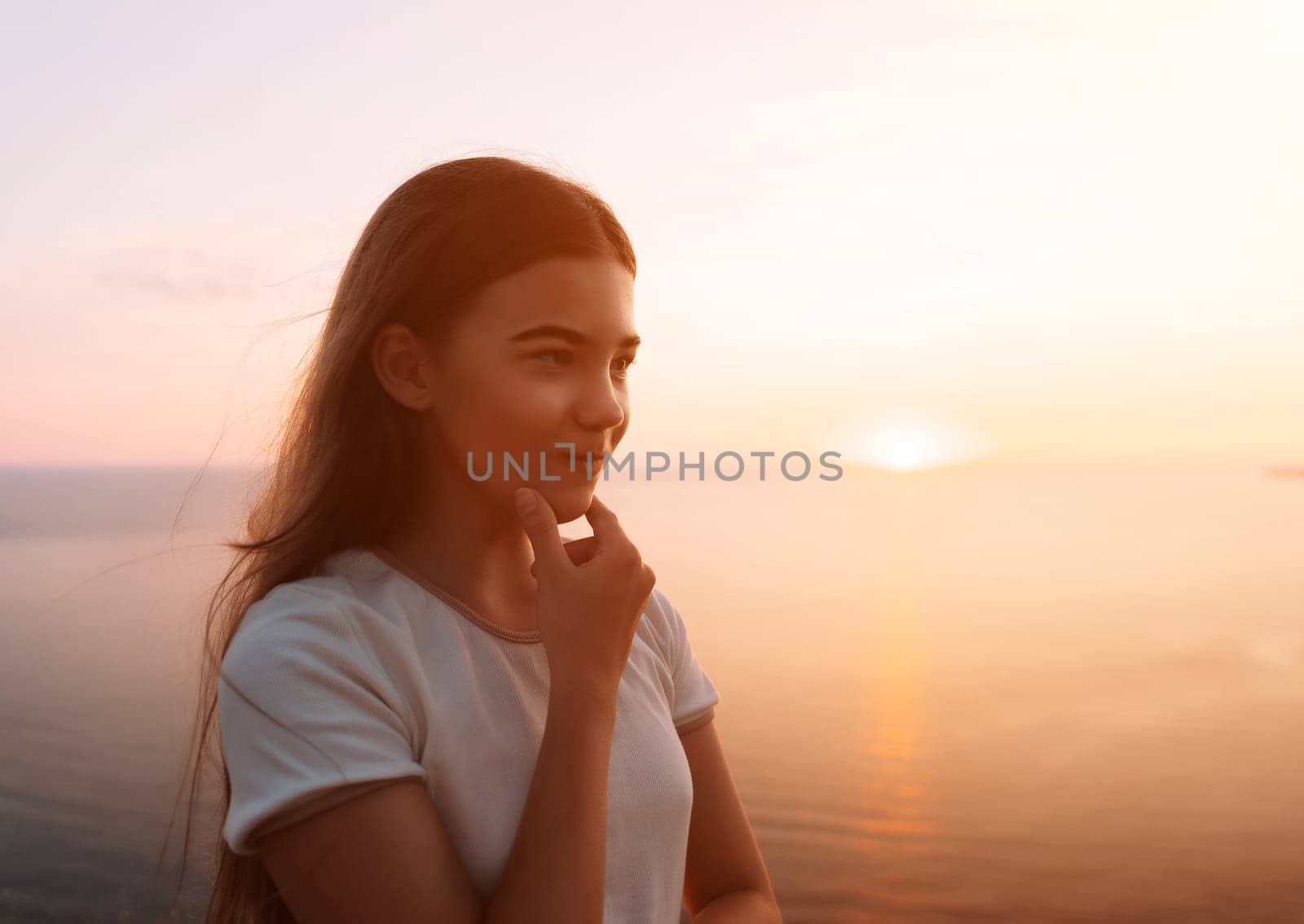A young girl with long brown hair stands by a lake at sunset, lost in thought, gazing at the horizon.