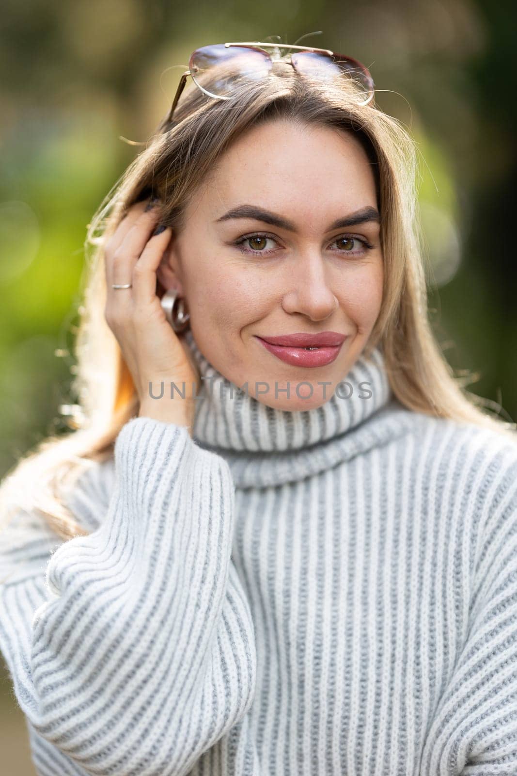 Attractive young woman with sunglasses on her head, wearing a white turtleneck sweater, smiling and touching her hair