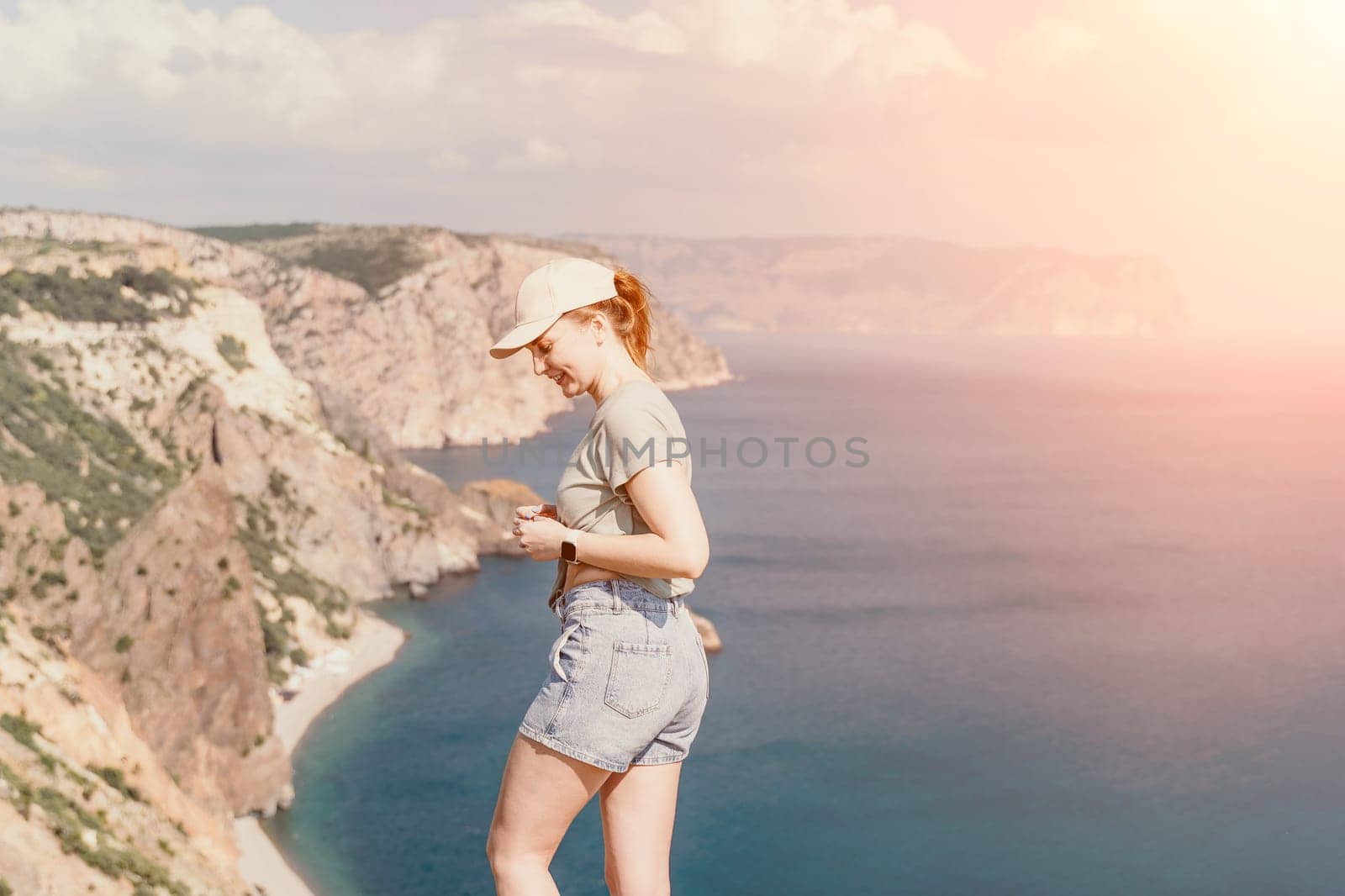 A woman wearing a cap and shorts stands on a cliff overlooking a vast ocean, bathed in warm sunlight.