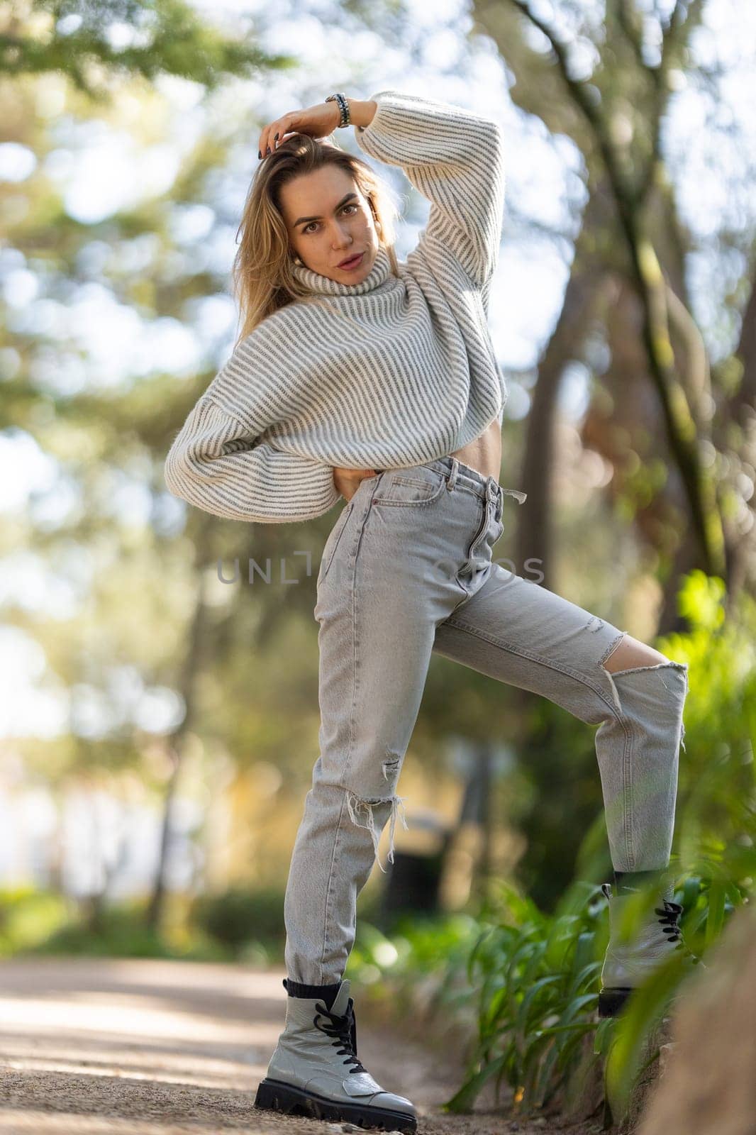 Young woman is posing in a park, touching her hair, wearing jeans and a sweater