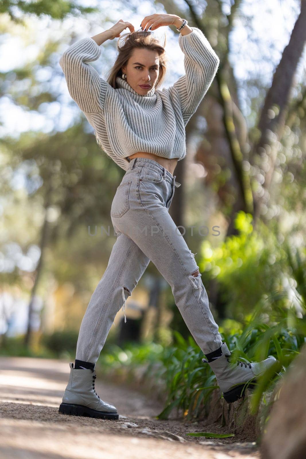 Young woman is standing on a path, holding her hair with her hands and wearing a wool sweater, jeans and boots