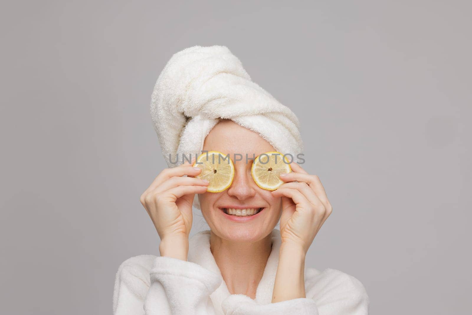A happy girl in a towel and robe holds two lemon slices in her hand, covering her eyes with an orange. Skin care and moisturizing concept