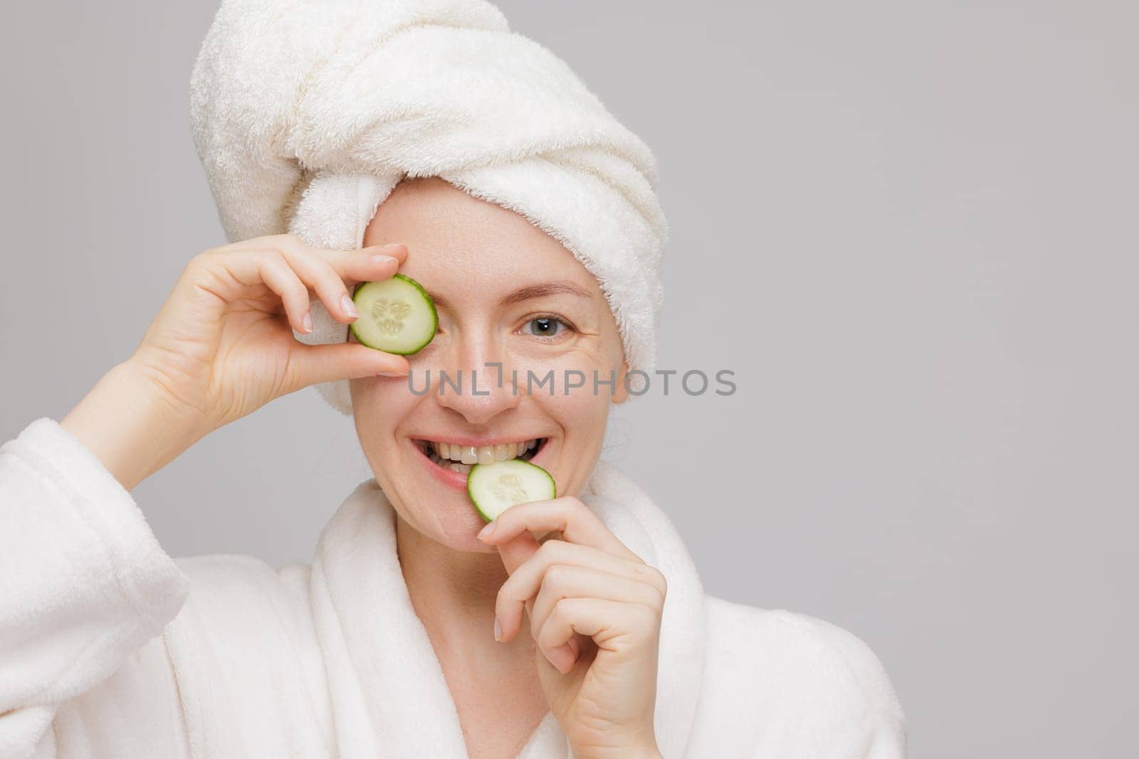 A woman in a white bathrobe and towel on her head holds cucumber slices over her eyes, smiling. The background is plain and light-colored, creating a spa-like atmosphere.
